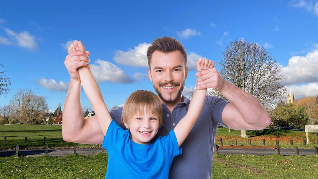 Father and young son celebrating with raised arms in Hurst Park's green space on a sunny day, with church spire visible in the background, representing the family connections fostered by HPRA community events.