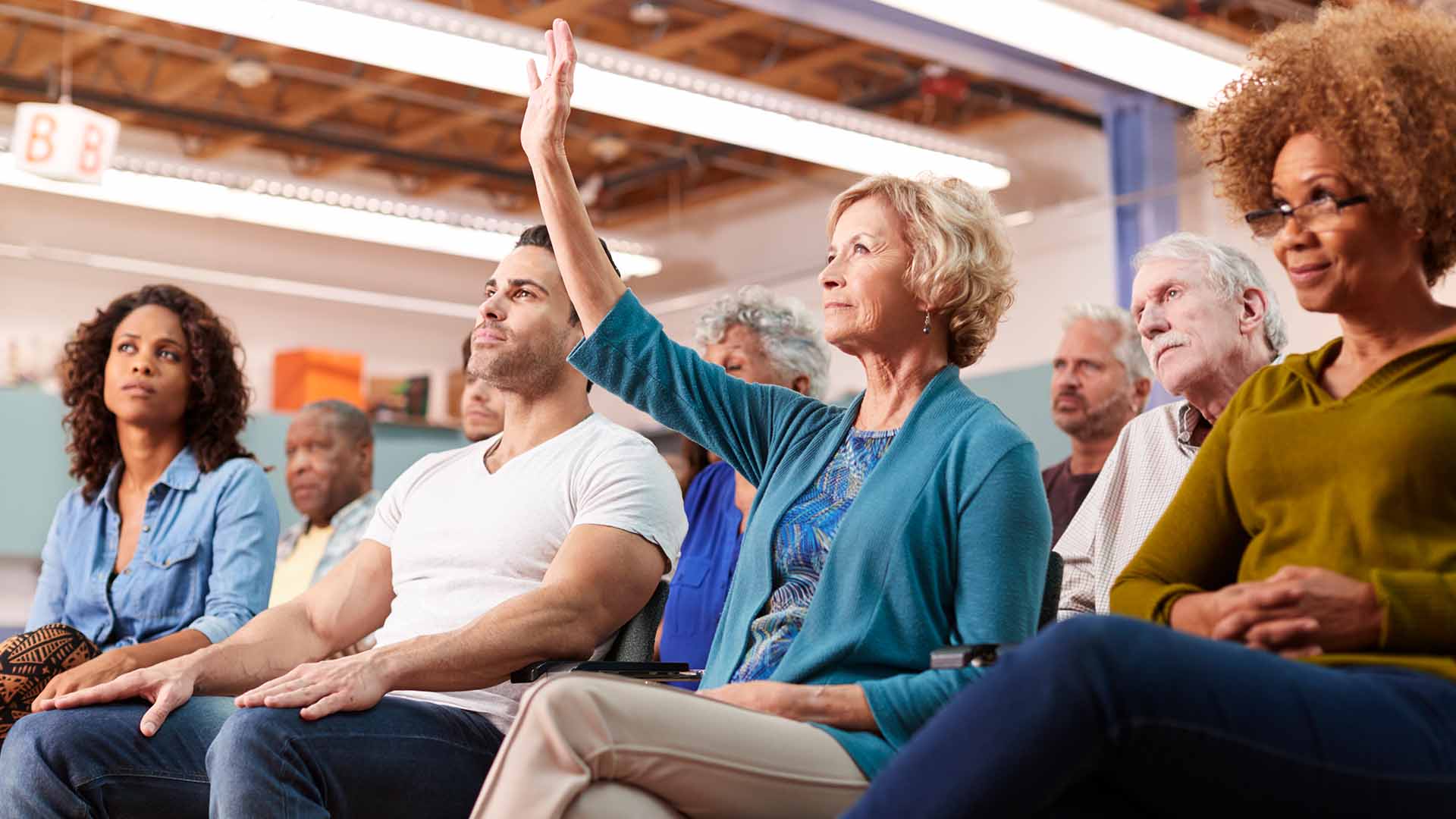 Diverse group of residents at a community meeting, with a woman in a blue cardigan raising her hand to ask a question, surrounded by engaged attendees in a community hall setting similar to where the HPRA Annual General Meeting will be held.