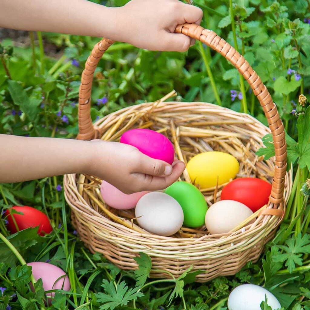 A child places a brightly colored Easter egg into a straw basket already filled with eggs, surrounded by green grass and spring flowers — symbolising the Easter Egg Hunt at Hurst Park.