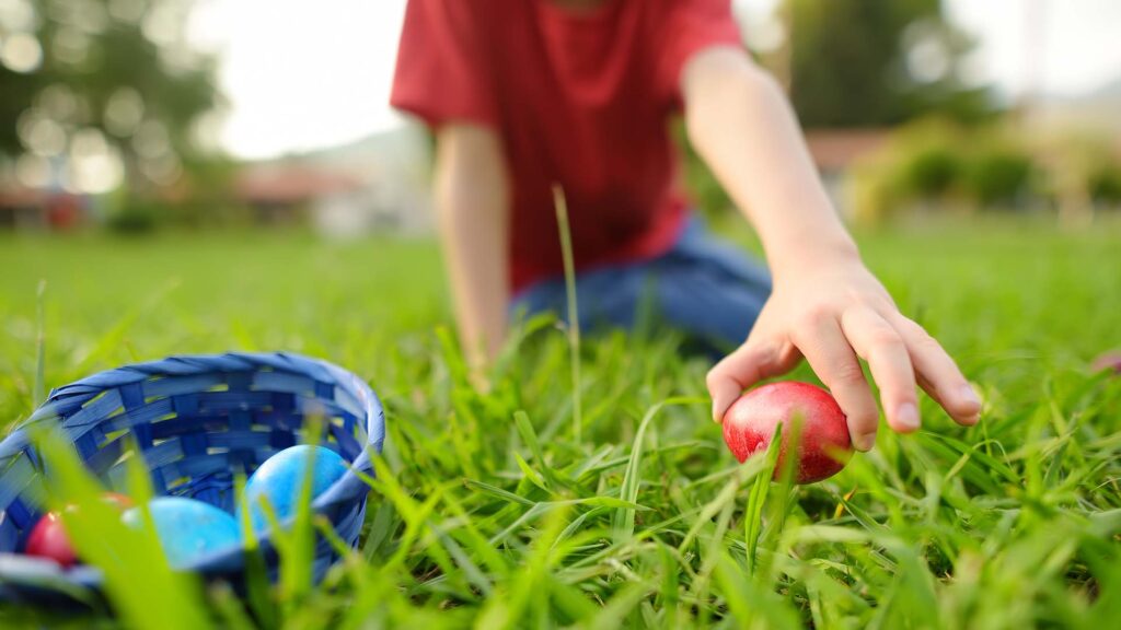 Child's hand picking up a red Easter egg from green grass with a blue basket containing colourful eggs nearby, illustrating Hurst Park's upcoming community Easter Egg Hunt event.