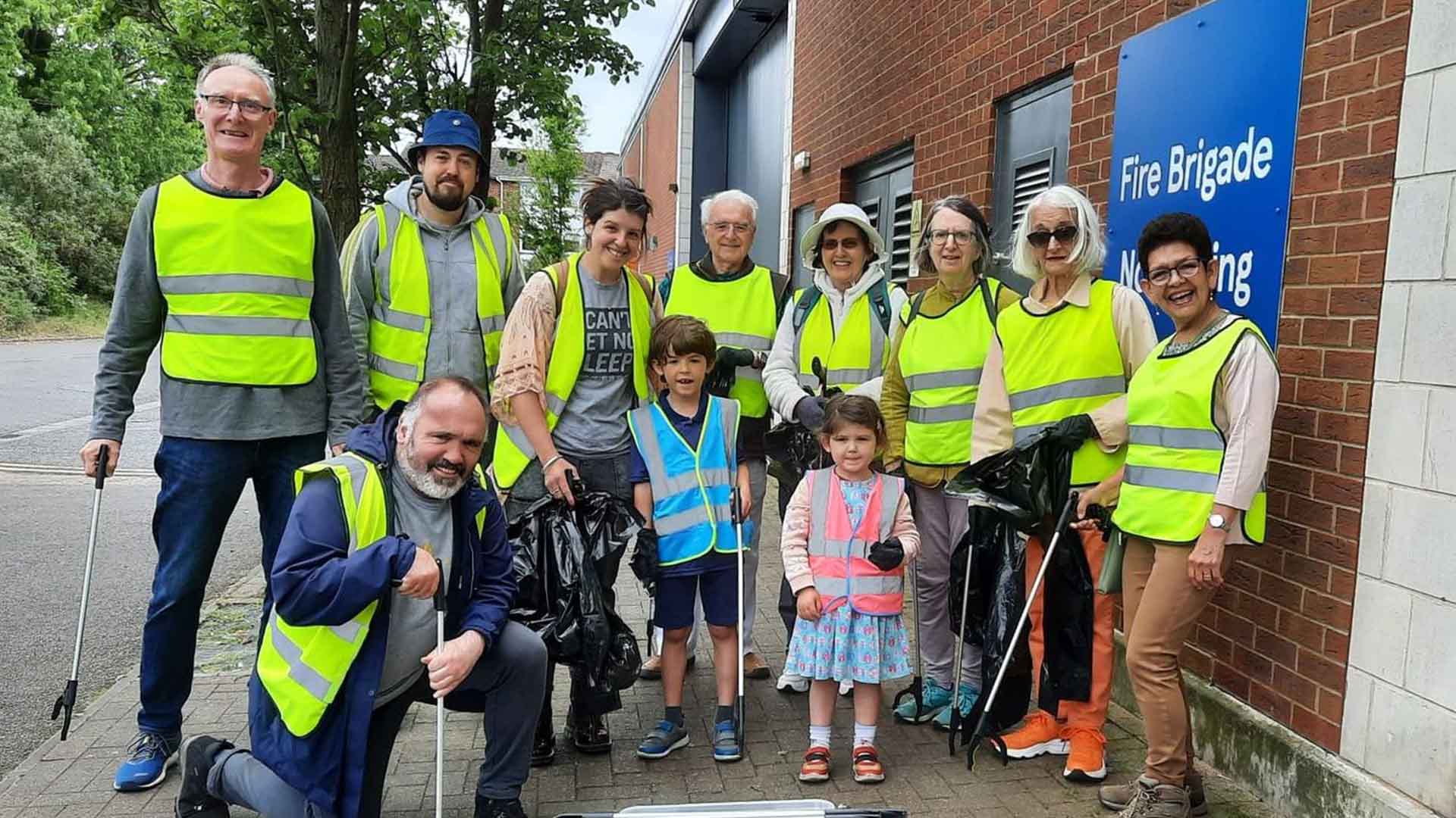 Group of community volunteers in high-visibility vests of various ages, including children, gathered outside of Tesco's Fire Brigade building with litter-picking tools and trash bags for a Hurst Park neighbourhood cleanup initiative