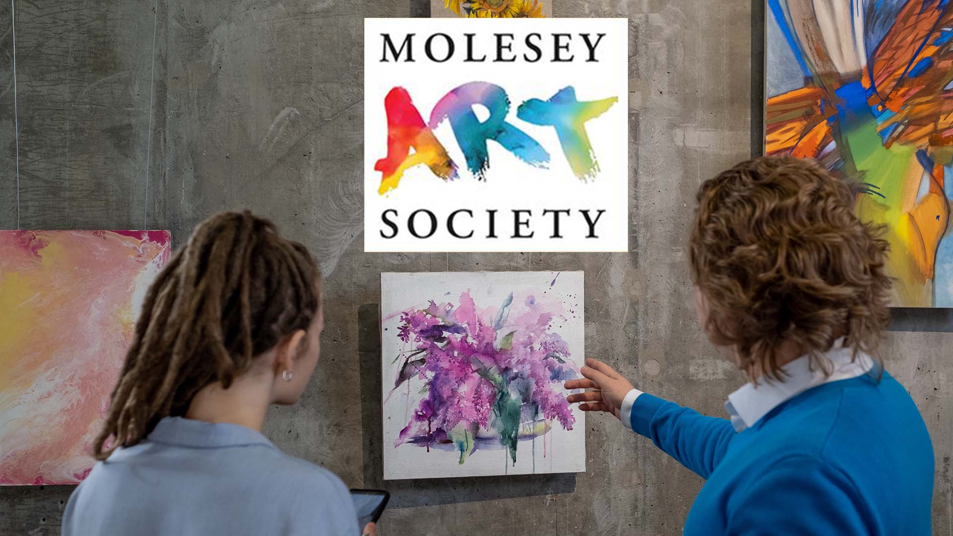 Two people viewing a watercolour artwork at the Molesey Art Society exhibition, with the colourful Society logo prominently displayed on the gallery wall showing 'ART' in various colours.