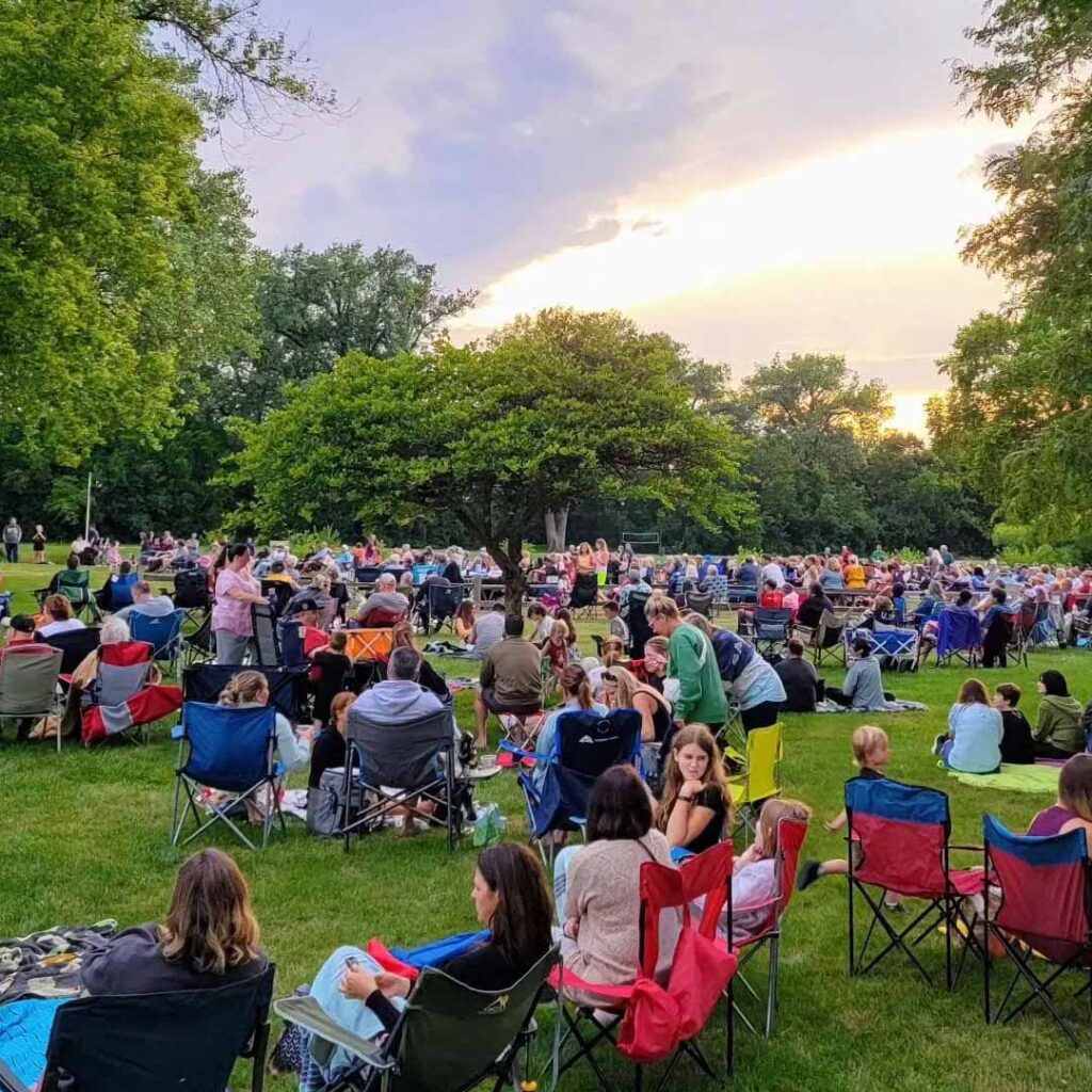 Crowd enjoying live music at Midsummer Music at the Marker in Hurst Park during sunset with chairs and trees.
