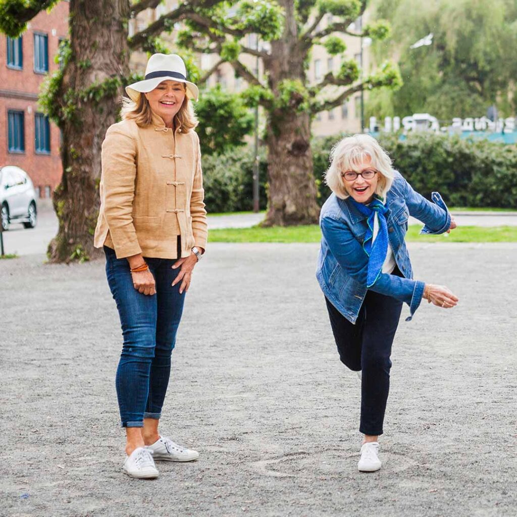 Two smiling women playing petanque at Hurst Park, one wearing a beige jacket and white hat, the other in a denim jacket demonstrating a throwing stance on the gravel terrain with trees and buildings in the background
