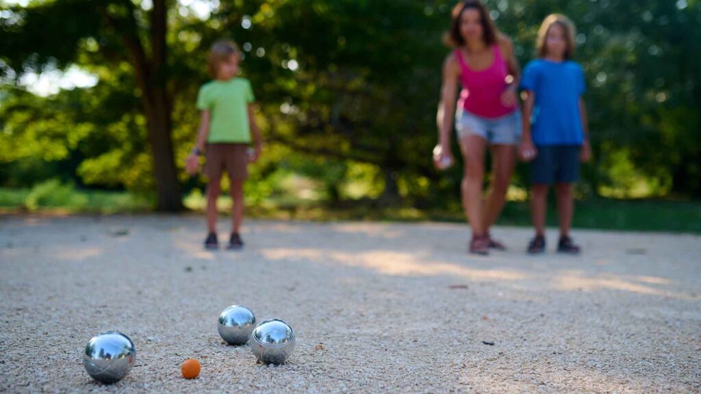 Close-up of silver petanque boules and a small orange jack on a gravel terrain, with blurred figures of children and an adult in the background enjoying a game at Hurst Park's petanque grounds.